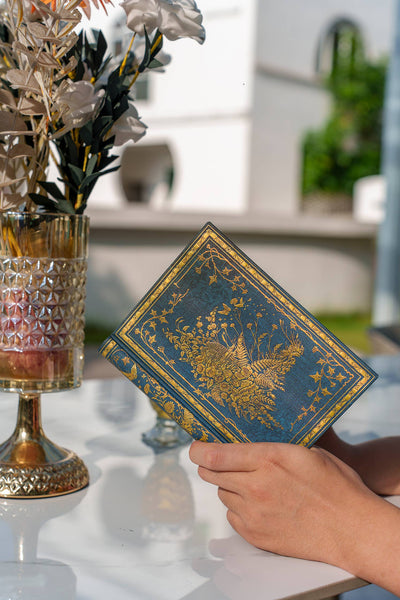 Hand holding a decorative card with floral design near a vase with flowers on a reflective surface.
