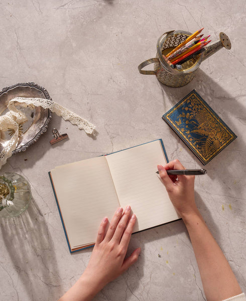 Person writing in an open notebook on a textured surface with a small container of pencils and decorative items.