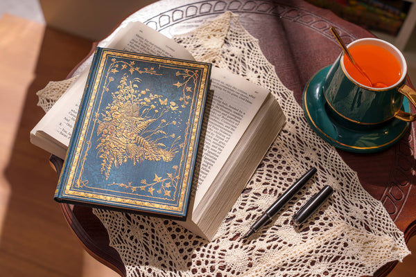 Open book with ornate cover on a lace tablecloth next to a cup of tea.